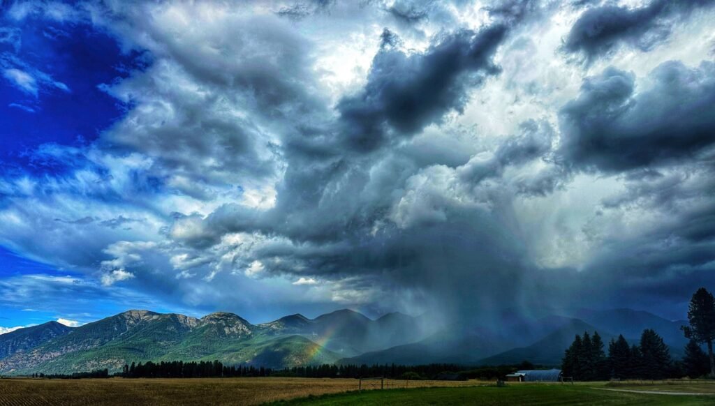 Dramatic storm clouds over green mountains, with a rain shaft and faint rainbow above the valley.