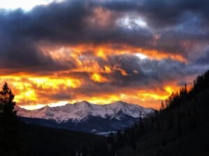 Fiery orange clouds above a snow-covered mountain range framed by dark forest at dusk.
