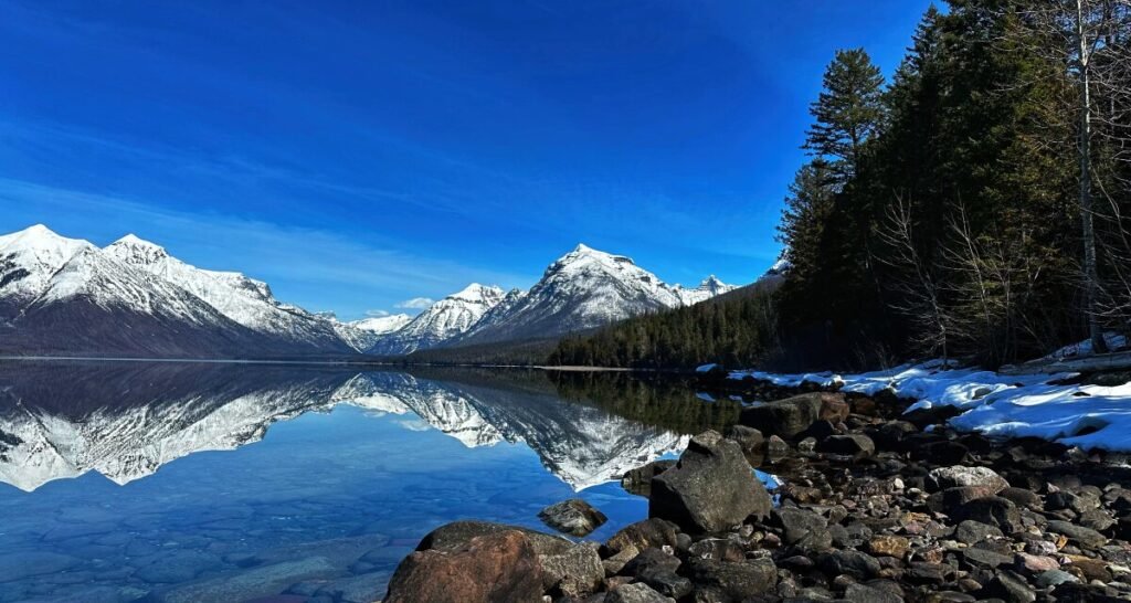 Snow-covered mountains reflected in a still lake beside a rocky shoreline and evergreen forest.