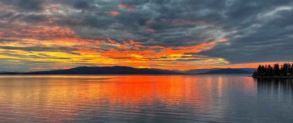 Fiery orange sunset clouds reflected across calm water, with dark mountains and a tree-lined shoreline.