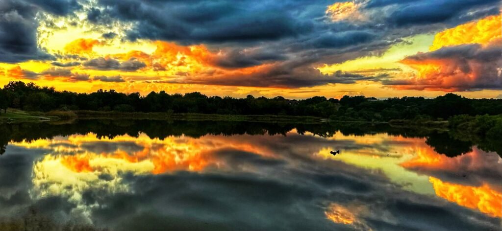 Fiery sunset clouds mirrored in still water above a dark tree-lined shoreline.
