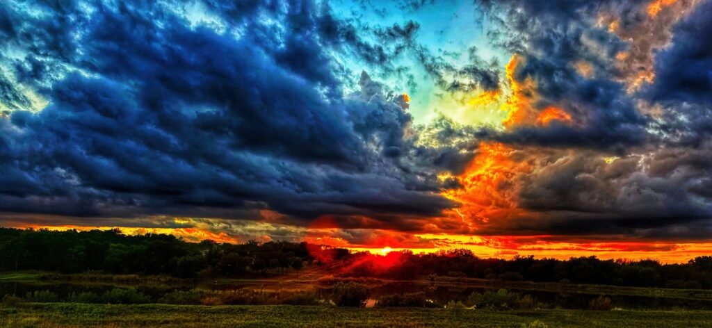 Intense orange sunset light breaks through dark storm clouds above fields and reflective water.